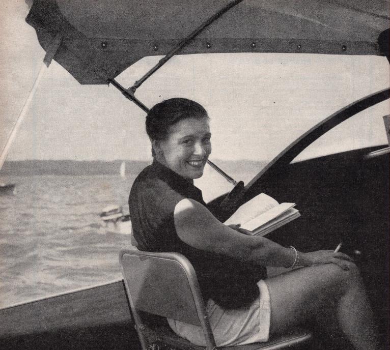 Virginia Kraft smiles over her shoulder, sitting on a boat, holding a cigarette in one hand and a book in the other. 