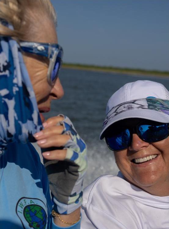 Two women sit on a boat chatting, wearing sunglasses, hair blowing in the wind.