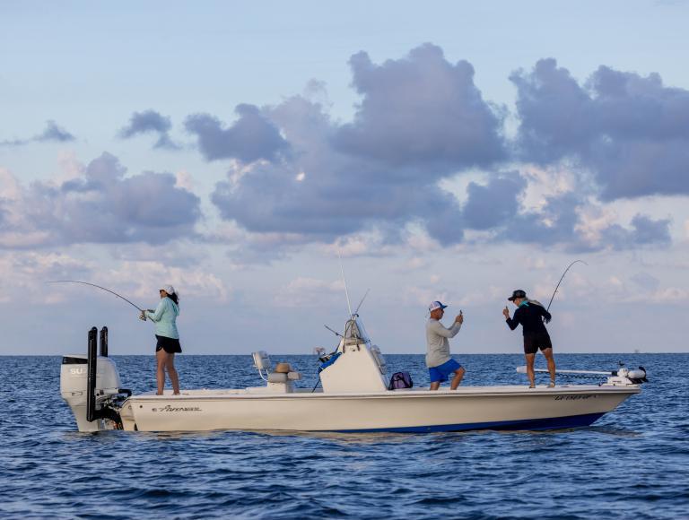 A man takes a photo of a woman giving a thumbs-up at the bow of a boat, while another woman fishes off the stern.