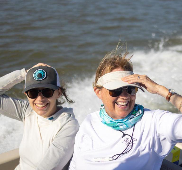 Two women smile on a boat, while holding onto their hats.