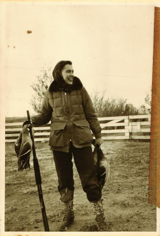 Virginia Kraft poses with fowl in each hand, smiling with a hunting rifle.