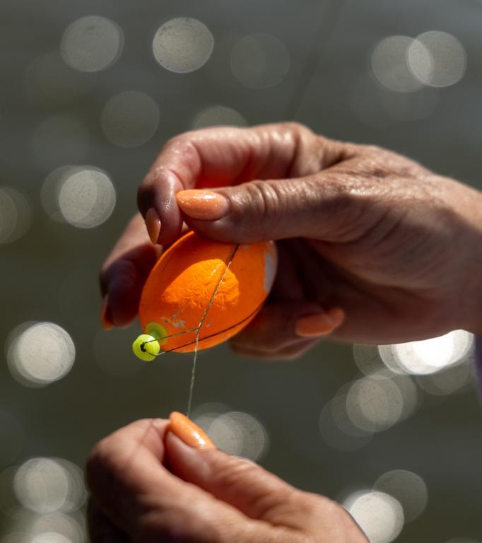 A woman&rsquo;s hand ties a bright orange bobber onto a fishing line.