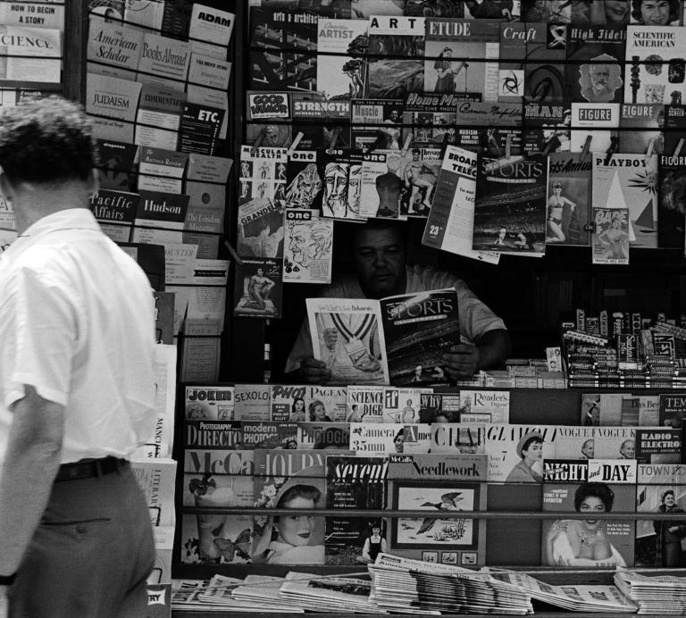 A news vendor looks through Sports Illustrated magazine in a stand surrounded by magazines and newspapers.