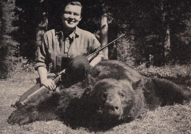 Virginia Kraft kneels next to a bear, holding her rifle and beaming. 