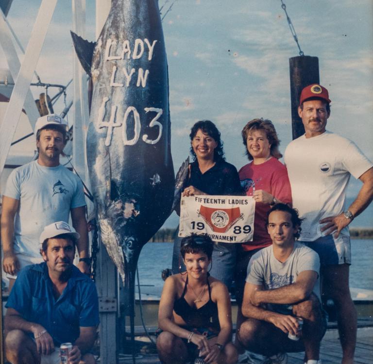 Men and women pose on a dock with a sign that reads Fifteenth Big Game Fishing Club, New Orleans, 1989.