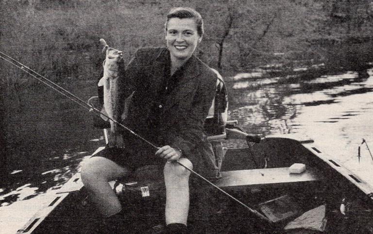 Virginia Kraft holds up a bass, smiling, in a small fishing boat.