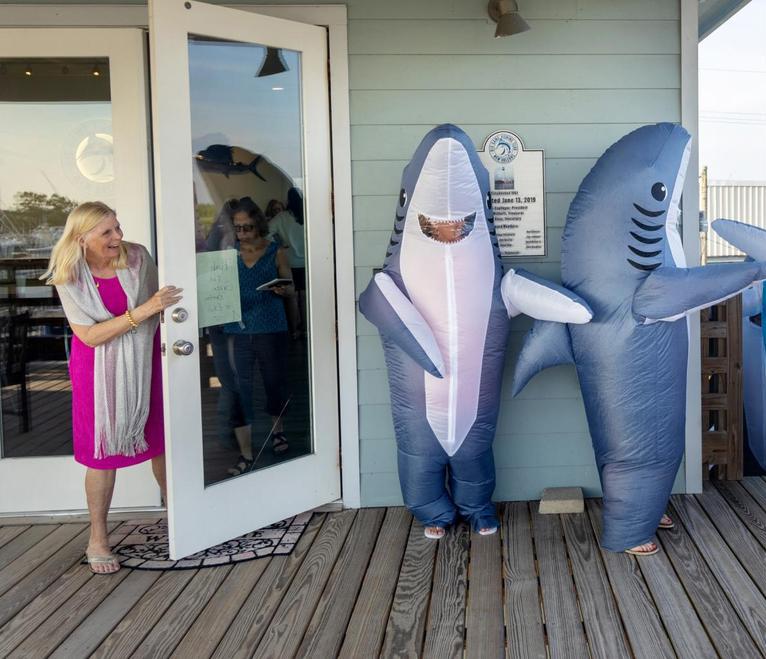 A woman laughs as she peeks around a door to an outdoor deck, with several women in inflatable shark costumes preparing for a dance.