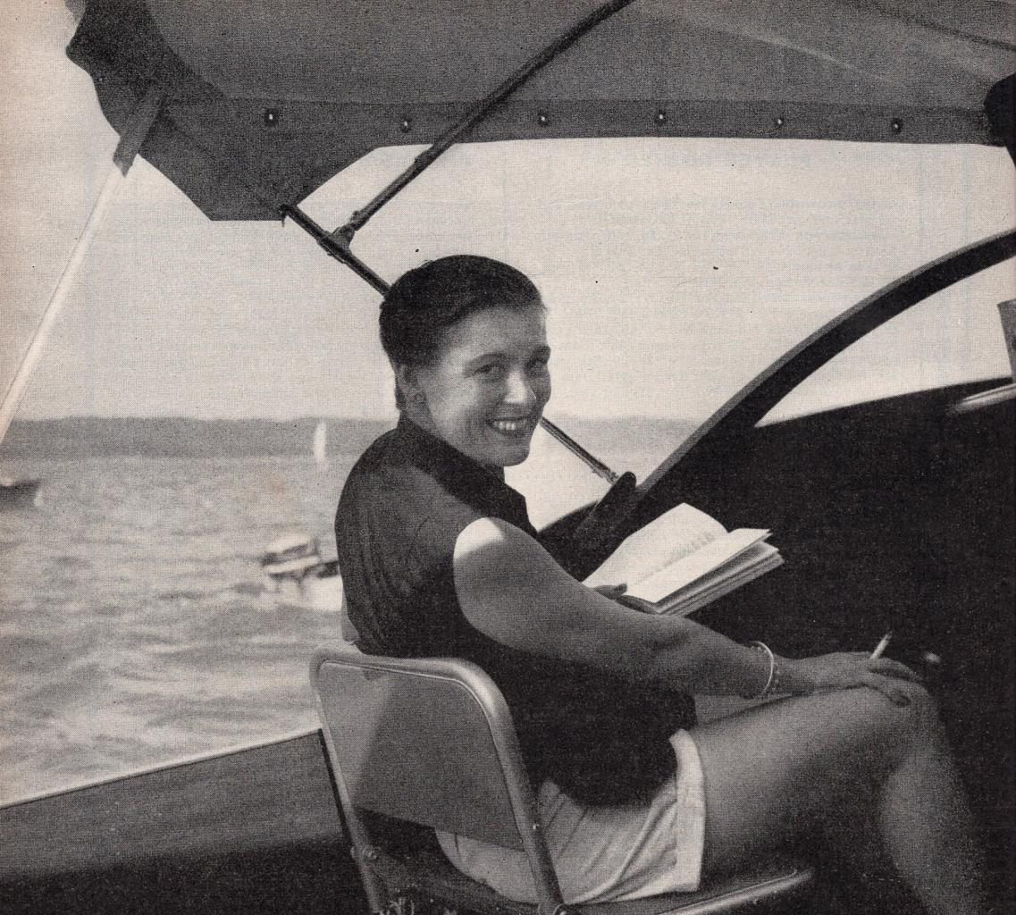 Virginia Kraft smiles over her shoulder, sitting on a boat, holding a cigarette in one hand and a book in the other. 