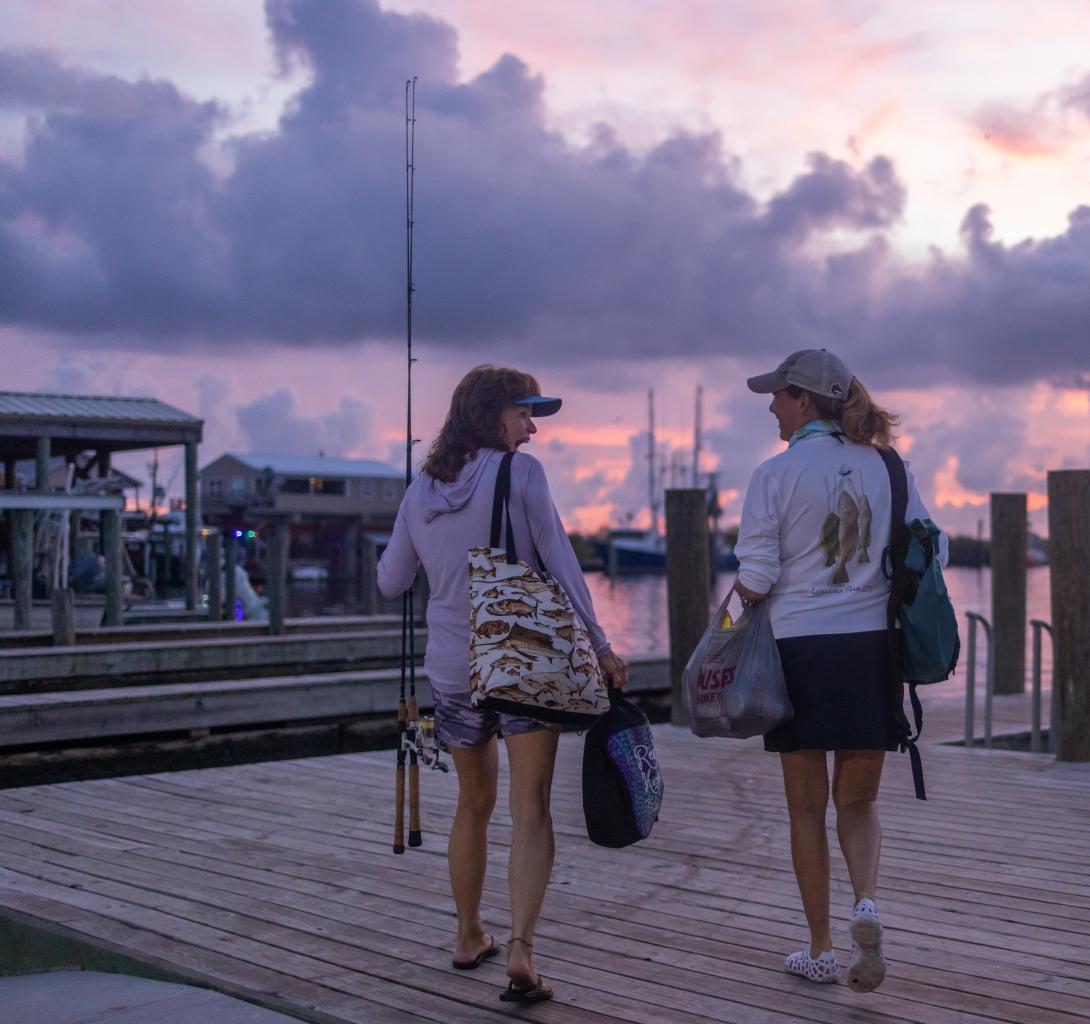 Two women, Leiza Fitzgerald and Connie O&rsquo;Day, walk down a dock, fishing poles in hand, to their boats before the 2023 Louisiana SLAM Tournament begins.