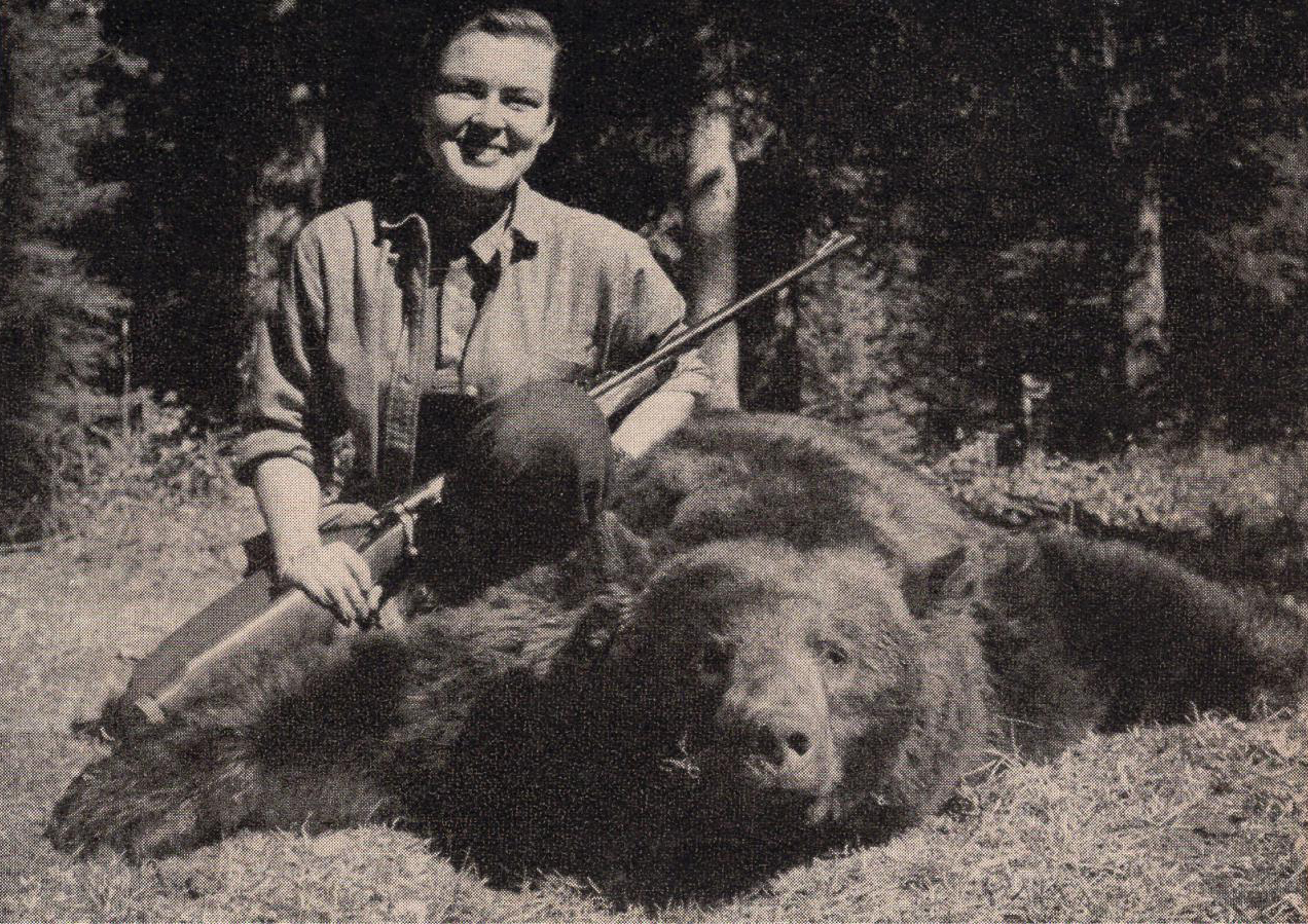 Virginia Kraft kneels next to a bear, holding her rifle and beaming. 